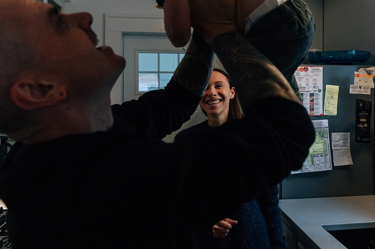 mom smiles through dad's arm while lifting toddler son in joy
