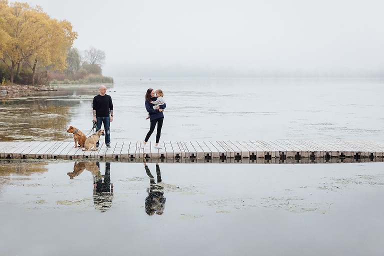 Dad holds leash of two dogs and mom holds daughter on a pier. They are reflected in the water on a foggy fall weekend