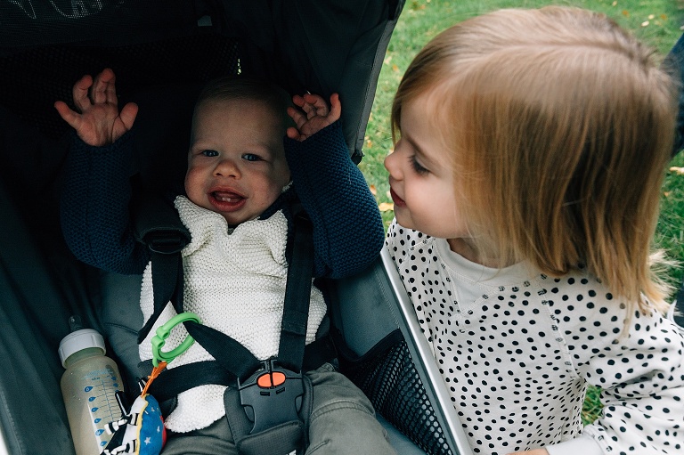 Girl smiles as she looks at toddler brother smiling in his stroller 