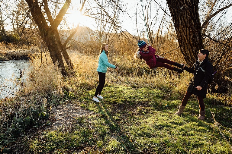 mom and sisters push sister on a wooden bench swing on a cold autumn day, sun flare in the background 