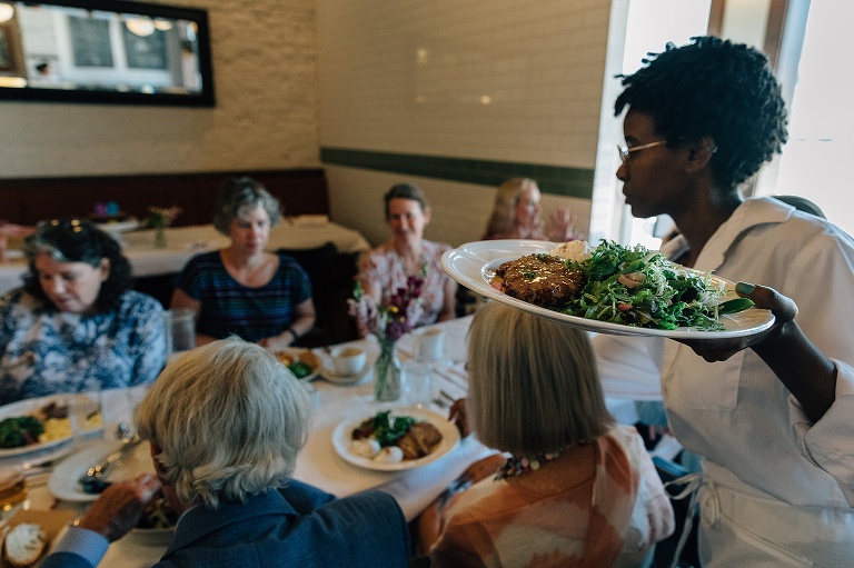 Waitress brings out a plate of food on a circular tray to a group of people at a long restaurant table