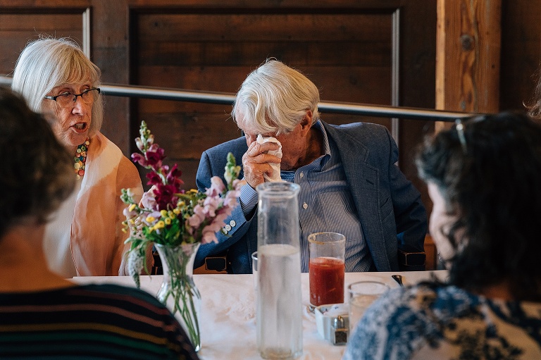 Husband of birthday girl dabs teary eyes with a white napkin at the end of a long table during an 80th birthday celebration