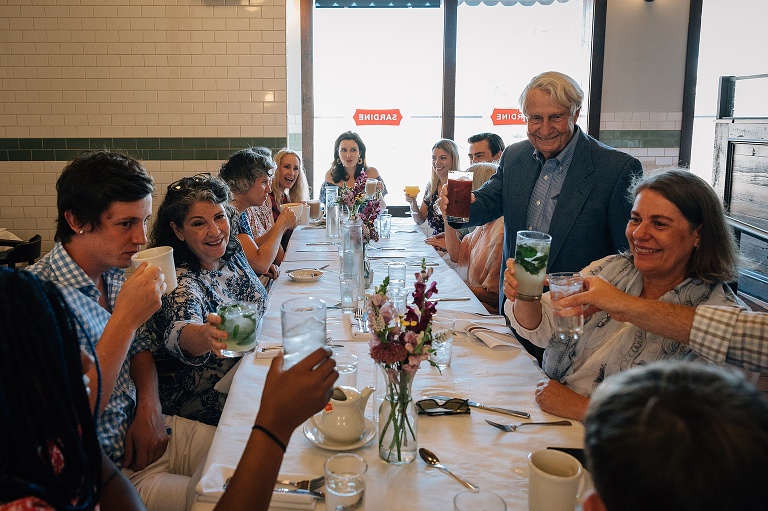 Man stands in the center of a long restaurant table to give a toast and cheers for an 80th birthday celebration