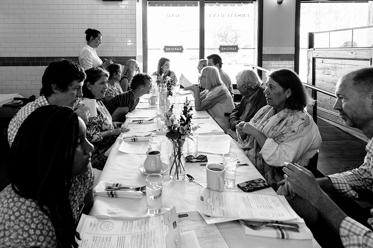 black and white; people at a long restaurant table making conversation for an 80th birthday celebration