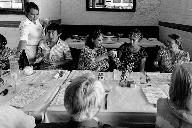 black and white; people at a long restaurant table making a "cheers" gesture with their cups