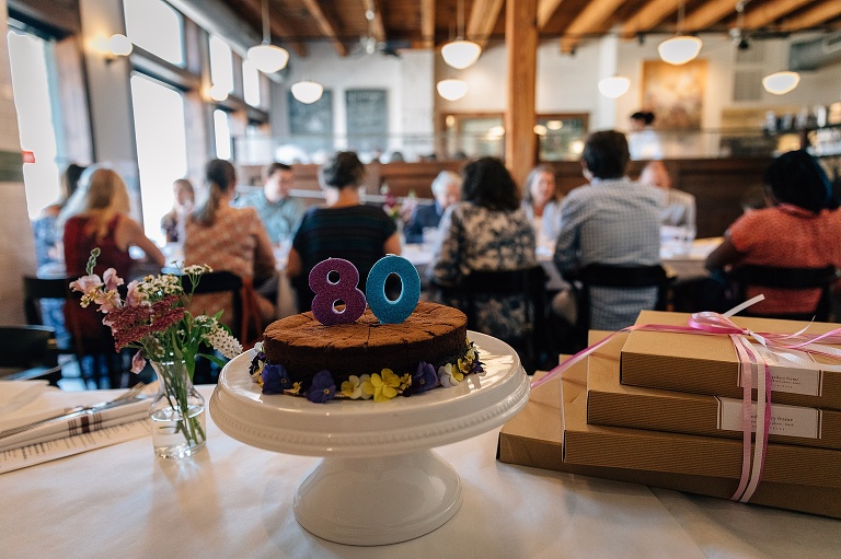 Focus on numeral 80 sitting on top of chocolate cake, presents on the side, family out of focus sitting at restaurant table