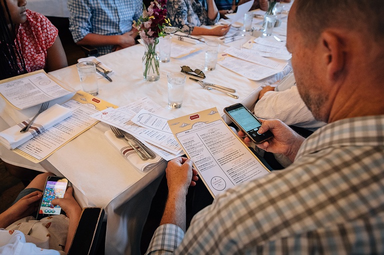  people at a long restaurant table examining the menu and covertly playing on their cellphone