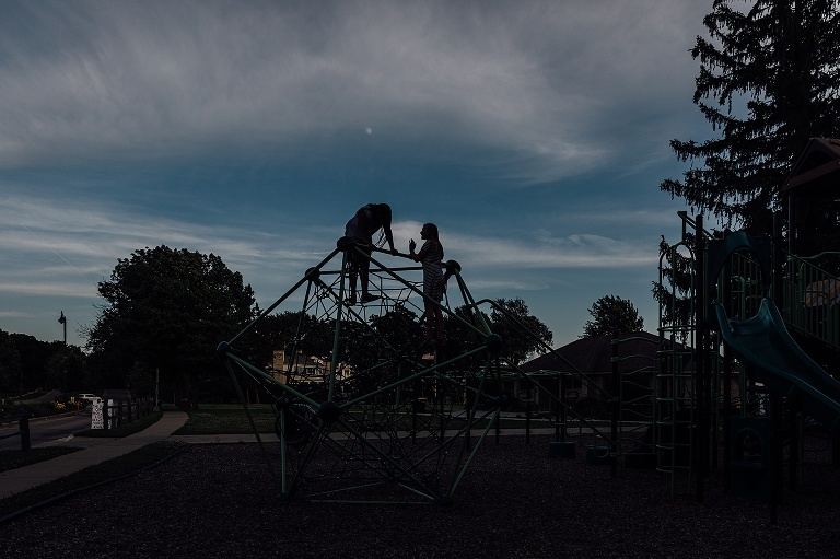 silhouette of two girls on a jungle gym playground equipment against a dusky sky with partial moon