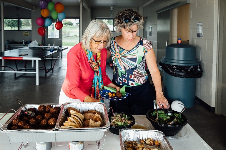 elderly mother and adult daughter point at prepared food buffet line at an event photography session
