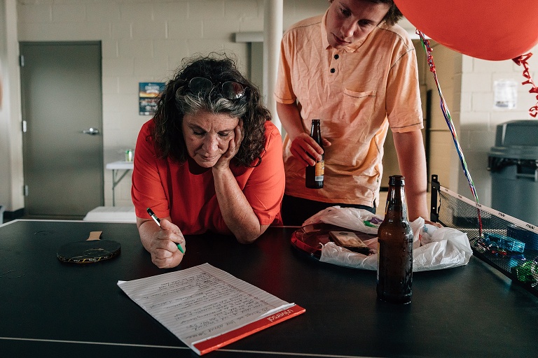 mother and son lean over a legal pad of paper with balloons and beer bottles in the frame