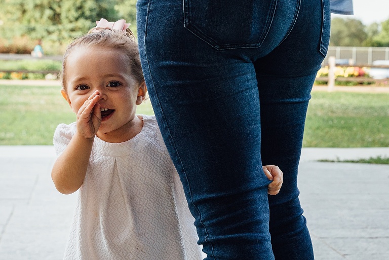 toddler cups a hand to side of her face and grabs on to her mother's jeans with the other