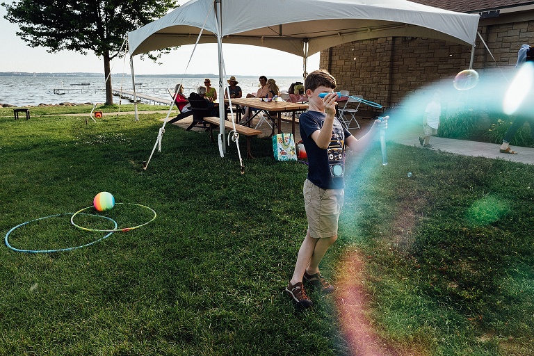 boy plays with giant bubbles at the beach while people gather under a tent in the background at an event photography session