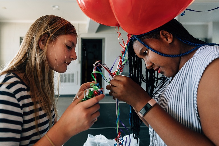 two girls untangle balloon ribbons together; red balloon above them