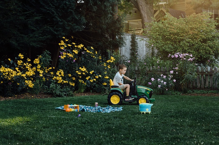 boy rides a toy tractor in his backyard. yard is filled with yellow flowers and evening sun. Backyard Summer Nights