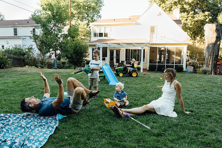 young boy points a toy leaf blower at his dad, while his mom and infant brother look on. Younger brother rides toy tractor in the background. 