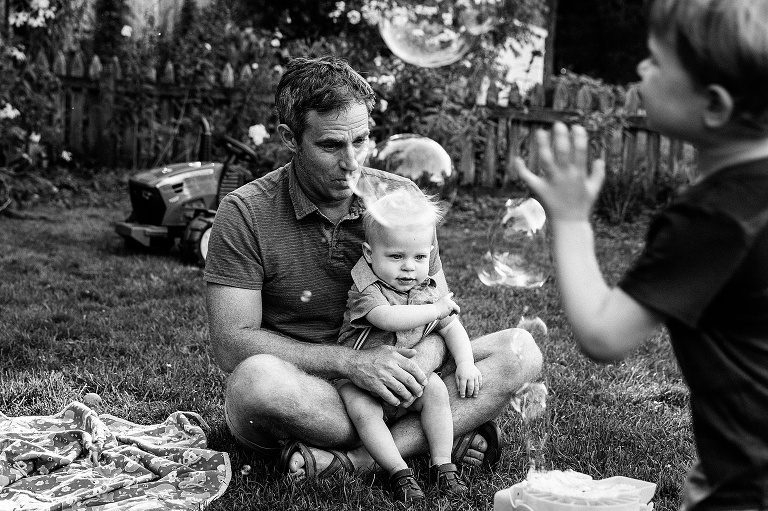 black and white. infant son sits on father's lap while a bubble machine fills the photo with bubbles