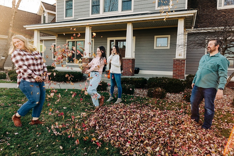 Teen daughters playfully toss fall leaves at their smiling mothers in front of their home