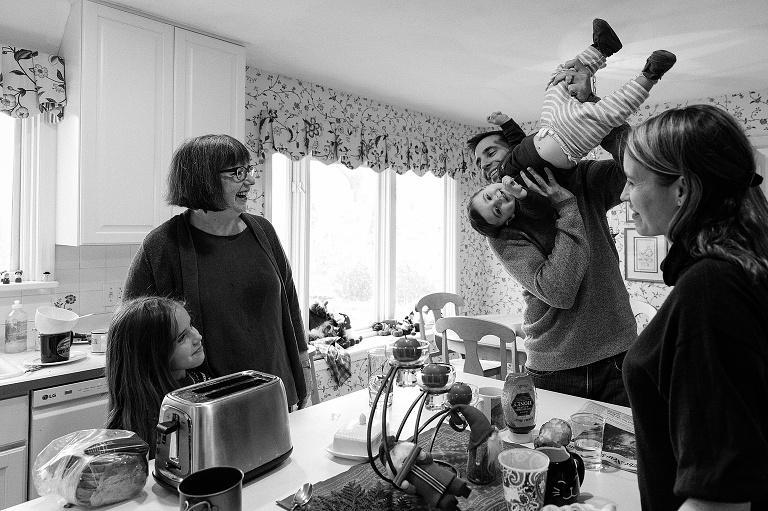 man holds toddler upside down in the kitchen, smiling, while mother, grandma, cousin watch on. black and white