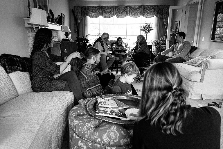 Large family scene in a living room. 6 adults and 3 kids visible. Ordinary scene of talking and reading together. 