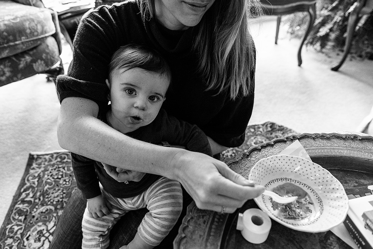 Mother holds infant while scooping food from a small bowl for baby. Baby is making eye contact with camera and mouth is open in anticipation of food. Black and white. 