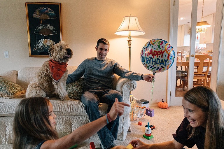 Girl and mother smile and gesture in the foreground while Dad holds Happy Birthday balloon in one hand and pets dog with the other hand.