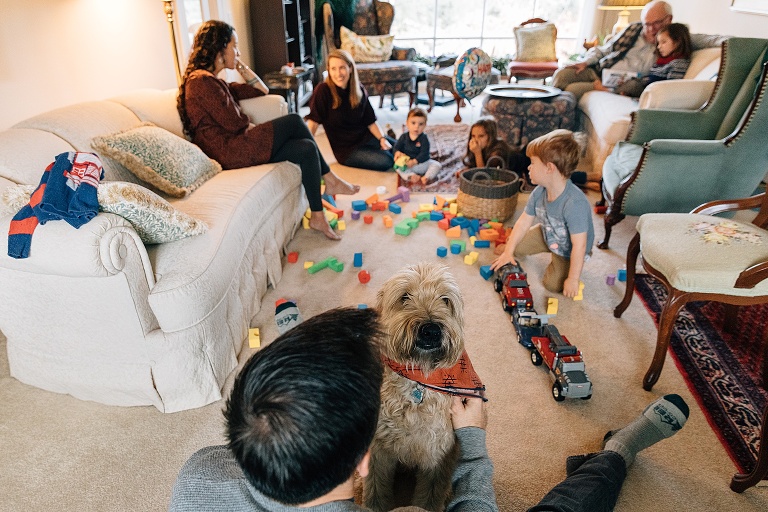 Focus on cute dog in a living room filled with 8 people, 4 of whom are children, and scattered toys.
