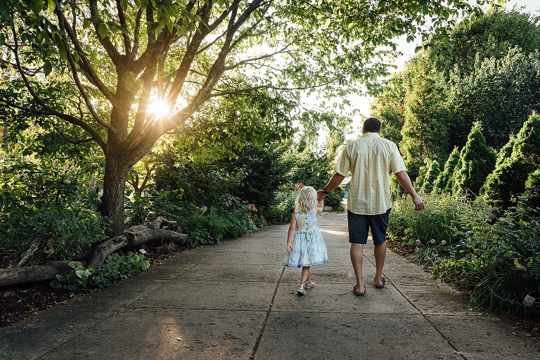 Dad holds daughter's hand as they walk toward sunshine coming through trees at Olbrich Gardens