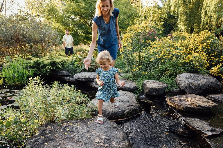 A mother reaches her hand out to young girl crossing a creek with stepping stones. Child smiling. Father watching and smiling from other side of the creek. 