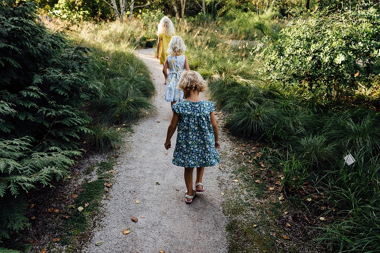 Three young girls in dresses explore a nature path that is sunlit up ahead.