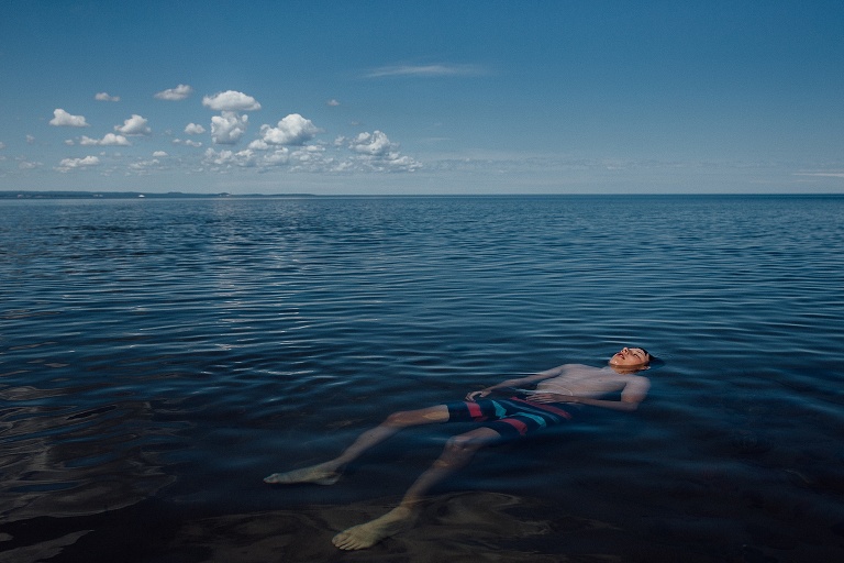 Boy lies in shallow like, floating. Blue lake, blue sky and white fluffy clouds are prominent.