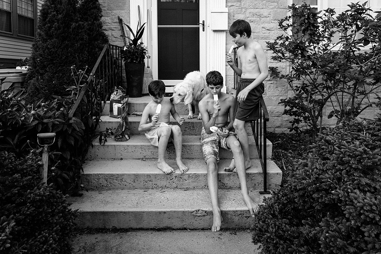 Three boys eat Popsicles on a front stoop while dog reaches for one of them over a boy's shoulder