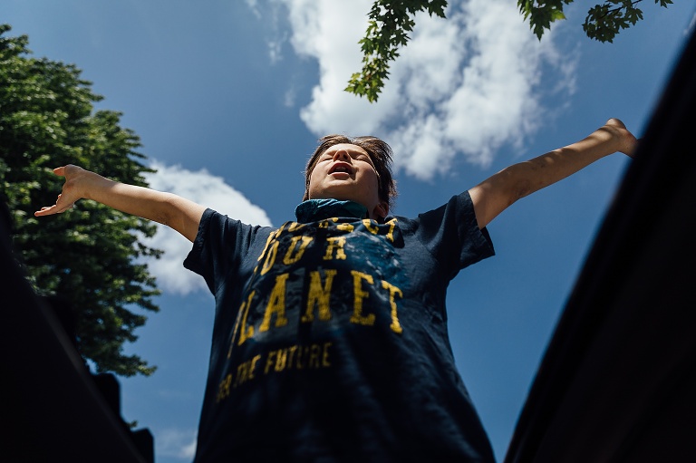 Boy outstretches arms with eyes closed against a blue sky and white clouds.