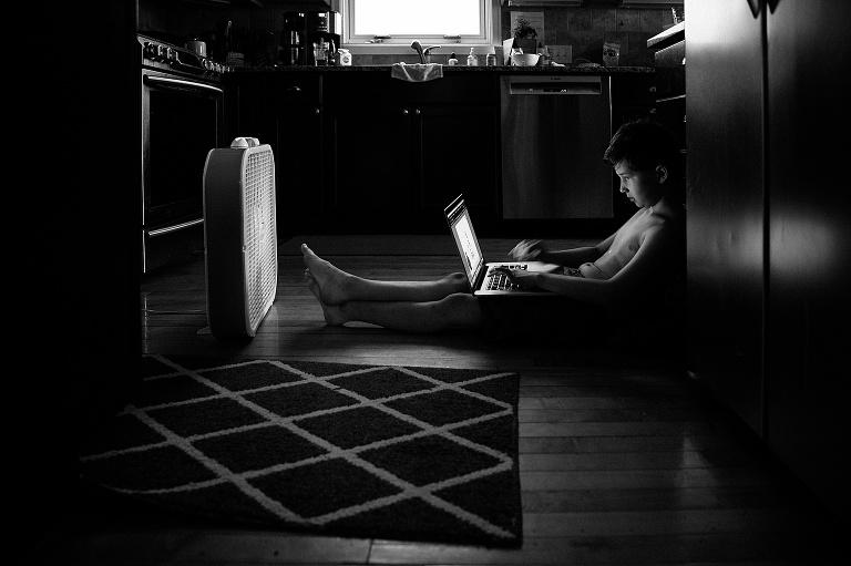 Boy sits on kitchen floor on the laptop while fan blows on him
