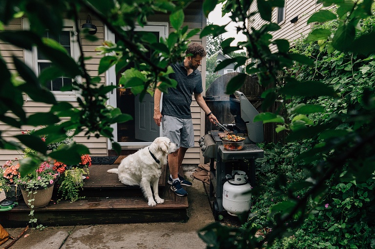 Man grills vegetables and steak while Golden Retriever sits and watches. 