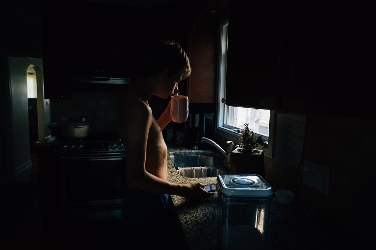 Teen stands at counter and drinks a cup of coffee in early morning light 
