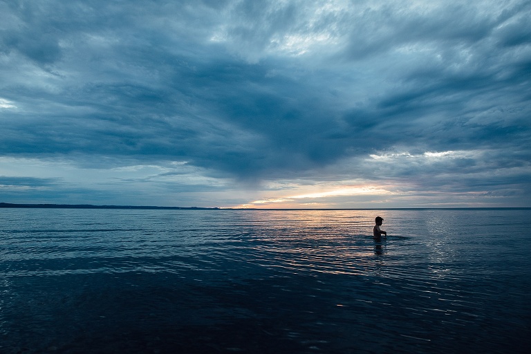 Child walks in a shallow lake at dusk with a sliver of warm light at horizon