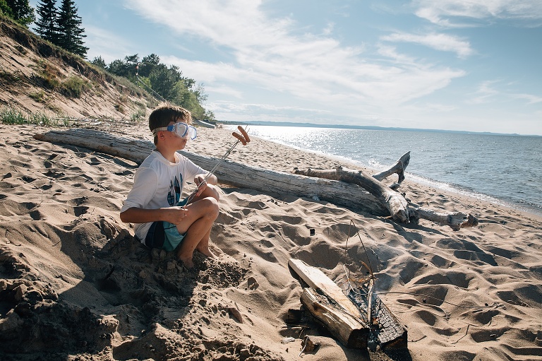 Boy wears scuba goggles on the beach over a smoky fire while roasting hot dogs