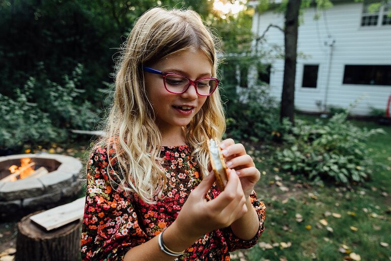 girl smiles at s'mores with fire pit and setting sun behind her