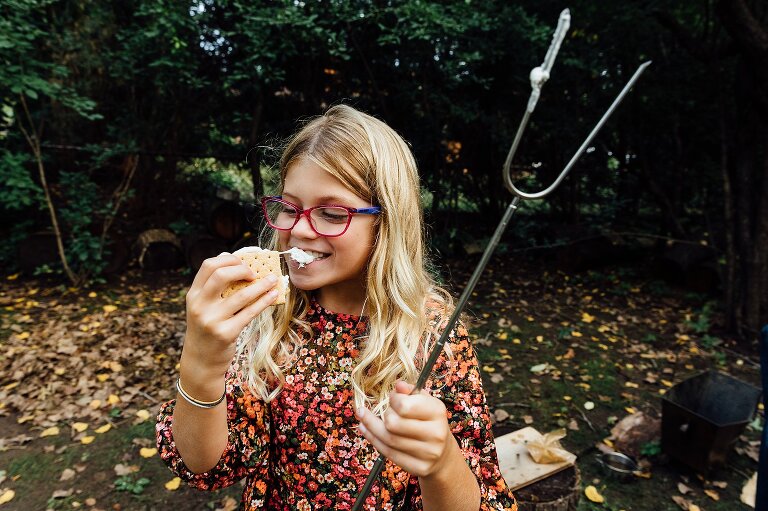 happy girl eats a s'more with stringy marshmallow, sticky skewer in her other hand