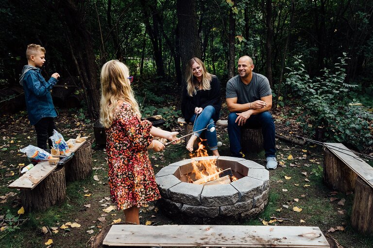 Family of four laugh and talk while making s'mores over open fire pit in the backyard