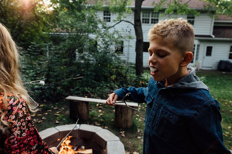 Boy pops marshmallow in his mouth. Setting sun and fire pit are present