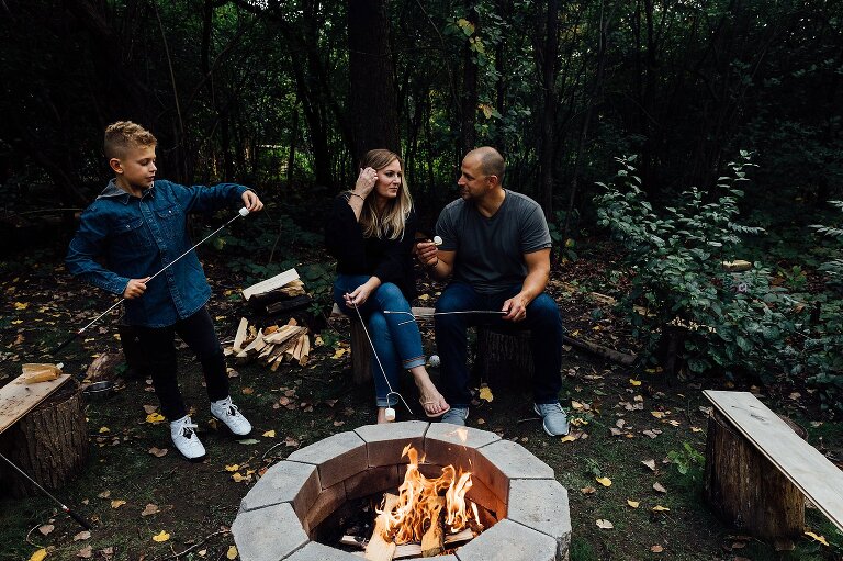 boy places marshmallow on end of skewer while mom and dad have conversation next to him. Fire pit in foreground.