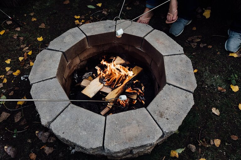 Fire pit fire in a circular pit in yard with yellow leaves; two marshmallows on skewers are placed over the fire