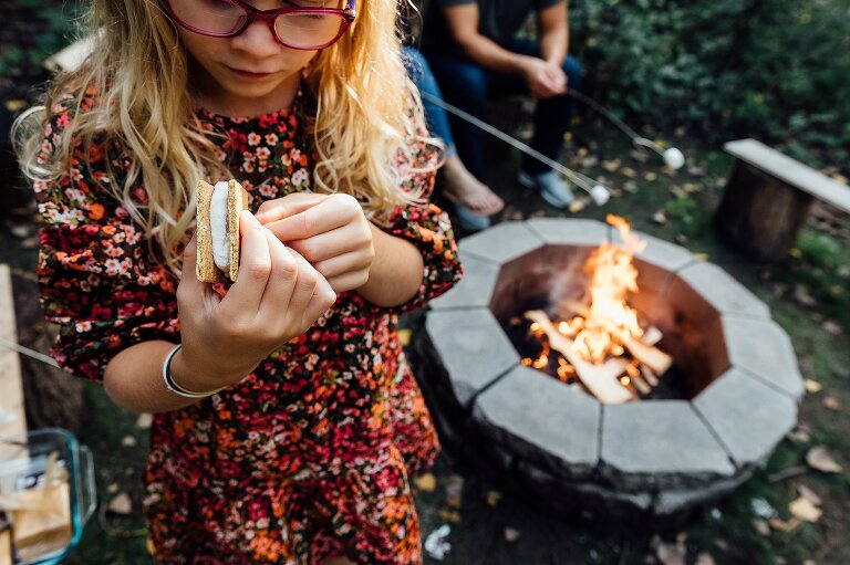 focus on girl's s'mores assembly while roaring fire in pit is in background