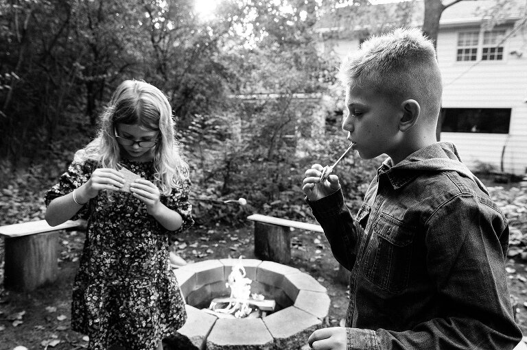 black and white. Boy bites into s'more and string of marshmallow trails out of his mouth; girl examines her s'more in background