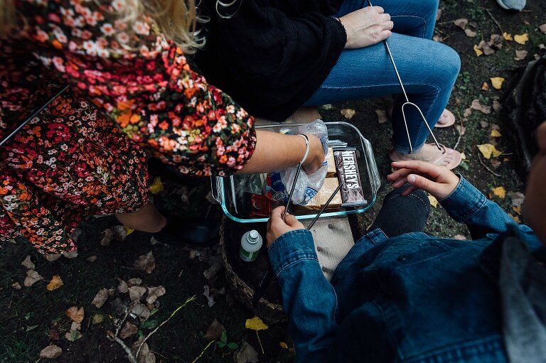 hands reach in marshmallow bag; chocolate bars, graham crackers and skewers are present