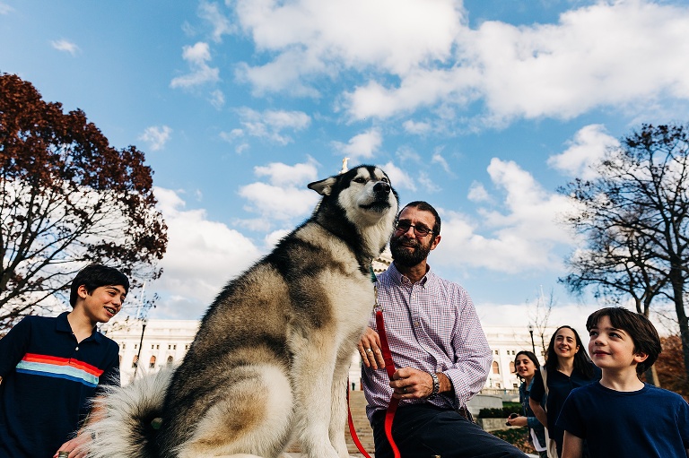 Dad smiles at dog who is centered in frame, rest of the family smiles from the edges of the frame 