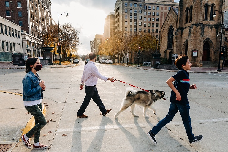 Man holds dog leash while teen boy and teen girl cross the urban street with setting sun on downtown Madison adventure 