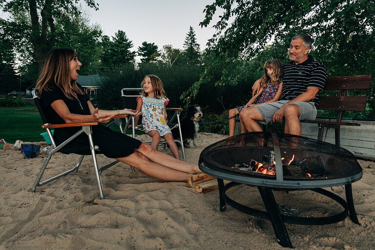 Family sits on the beach by fire pit. 
