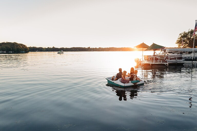 Family of four and dog ride paddle boat on lake in the sunset.